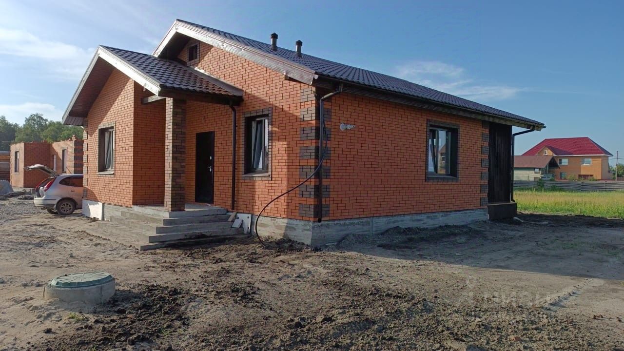 Дом выполнен в современном архитектурном стиле с кирпичной кладкой оранжевого цвета. Главной особенностью является симметричная форма с двускатной крышей, покрытой черепицей. Входная группа оформлена лестницей, ведущей к двери, с боковыми окнами, обеспечивающими естественное освещение. Стены отделаны кирпичом, дополненным декоративными элементами. Цветовая гамма включает теплые оттенки, создающие уютную атмосферу. Окна имеют черные рамки, что контрастирует с основным цветом здания. Внешний вид подчеркивает прочность конструкции и современный дизайн.