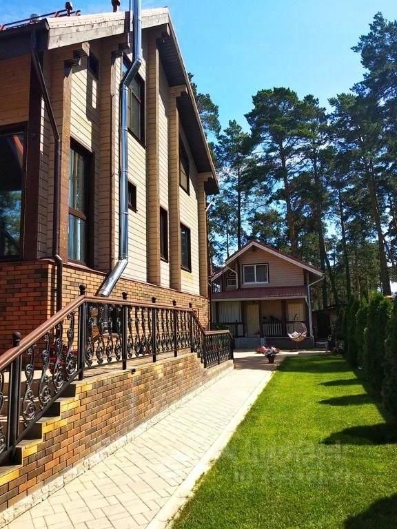 Архитектурный стиль здания характеризуется современными элементами с использованием дерева и кирпича. Видна лестница с кованым ограждением, ведущая к входу. Структурные особенности включают большие окна, обеспечивающие естественное освещение. На переднем плане расположена ухоженная зеленая территория с газоном и дорожкой. Основные цвета фасада - теплые оттенки дерева и кирпича, что создает уютную атмосферу. В задней части видно небольшое строение, вероятно, гостевой домик, с крышей, покрытой черепицей.