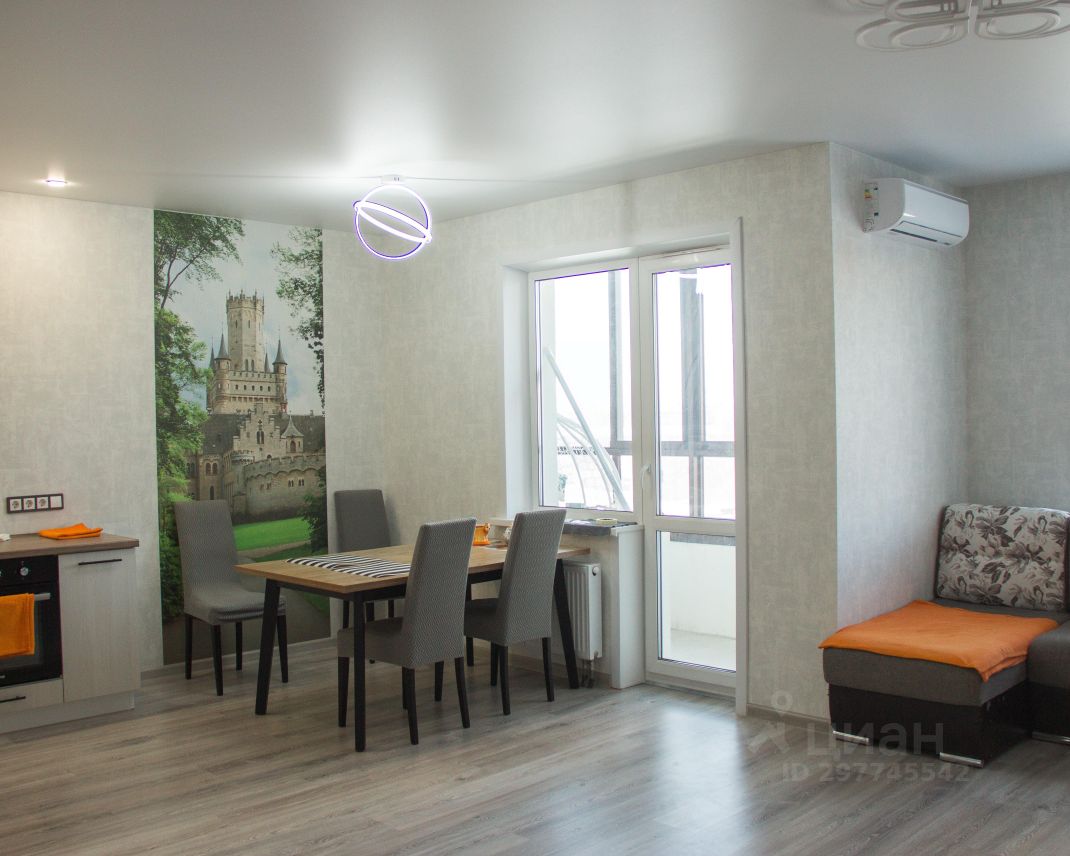 Современный интерьер с элементами минимализма. Стены оклеены светлыми обоями с текстурой, одна из стен оформлена фотопечатью с изображением замка на фоне зелени. На полу уложен ламинат светлого оттенка. В центре комнаты расположен обеденный стол из дерева с черными ножками, окруженный серыми стульями. В углу находится угловой диван с цветочной обивкой и оранжевым подушкой. Освещение представлено оригинальной люстрой в виде окружности, а также встроенными светильниками. Окно обеспечивает естественное освещение, создавая уютную атмосферу.