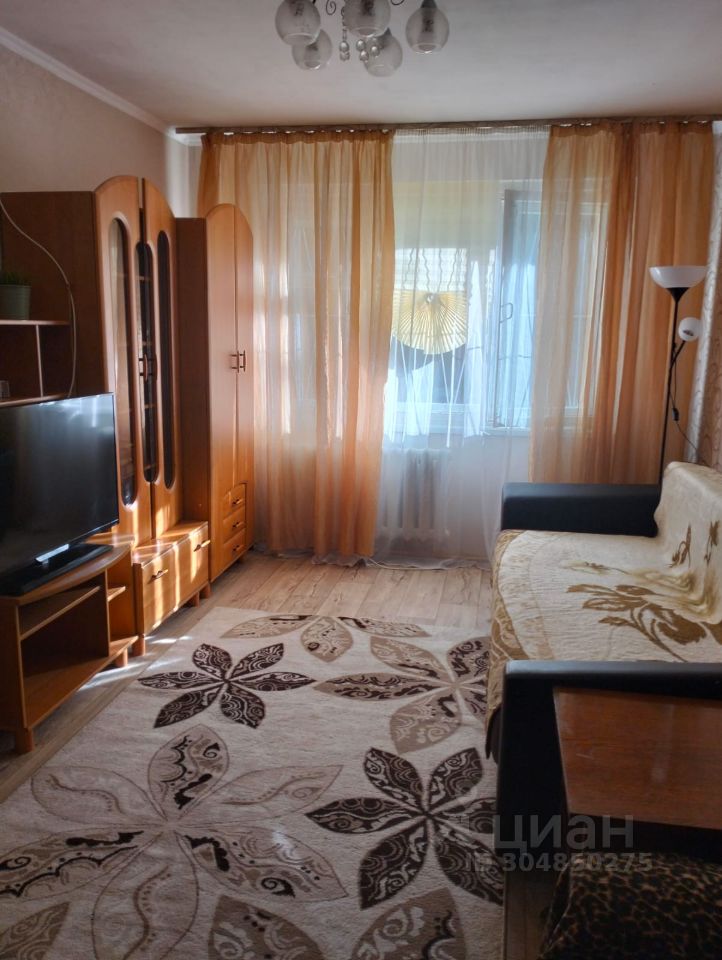 В комнате представлены элементы современного дизайна с использованием светлых и теплых тонов. Стены окрашены в нейтральный цвет, а пол покрыт светлым ламинатом. Мебель включает в себя деревянный шкаф с зеркальными дверцами и телевизор на тумбе. Угловой диван с текстильным покрытием и декоративными подушками создает уютную атмосферу. Окна обрамлены полупрозрачными шторами, которые пропускают мягкий свет. На полу расположен ковер с цветочным узором, добавляющий текстуру и уют. Освещение обеспечивается стильной люстрой и настольной лампой.