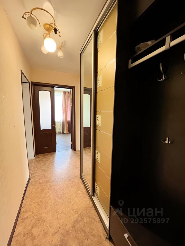 В коридоре виден современный стиль с элементами минимализма. Стены окрашены в светлые тона, пол покрыт ламинатом. Слева расположен шкаф-купе с зеркальными дверцами, что визуально расширяет пространство. Справа находится открытая вешалка для верхней одежды. Освещение обеспечивается стильной потолочной люстрой с тремя лампами. В конце коридора видна дверь, ведущая в светлую комнату с окнами, оформленными легкими шторами. Общая атмосфера пространства уютная и функциональная, что делает его удобным для повседневного использования.