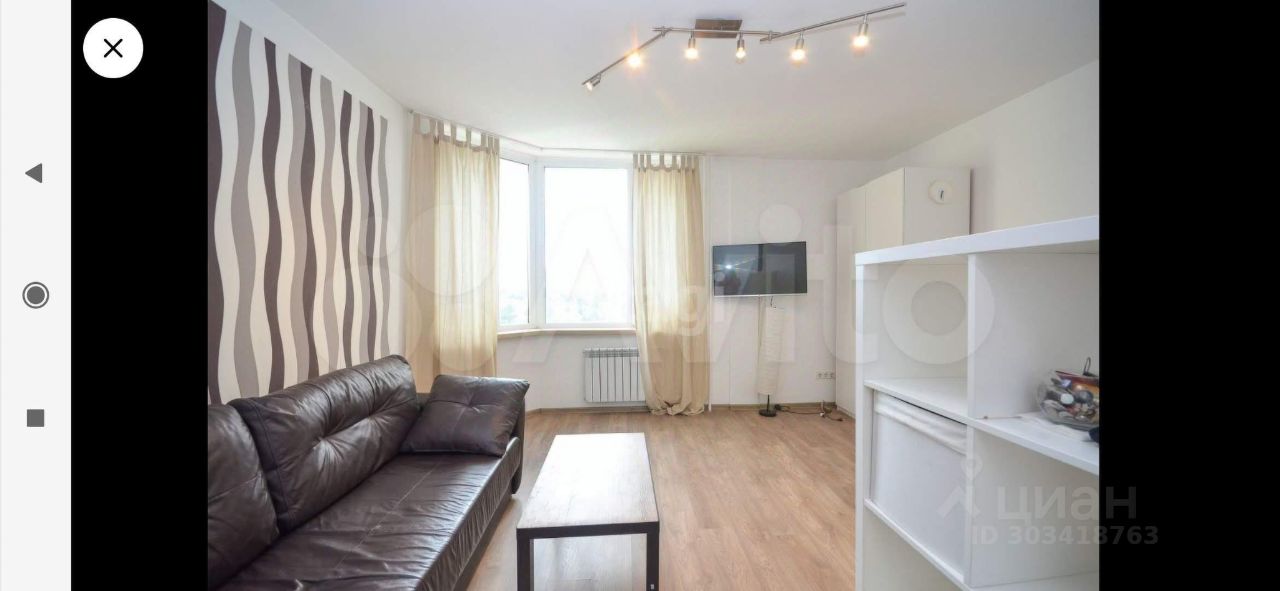 В комнате преобладает современный стиль с акцентом на минимализм. Стены оформлены в светлых тонах с волнообразным узором в коричневых оттенках. Просторное окно с легкими занавесками обеспечивает естественное освещение. В комнате находится удобный кожаный диван и деревянный столик, создающие уютную атмосферу. На стене размещён телевизор, а рядом стоит белый шкаф, который добавляет функциональности. Полы выполнены из светлого дерева, что визуально увеличивает пространство и придаёт ему современный вид.