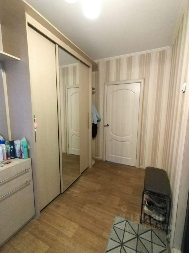 В прихожей присутствует современный стиль с элементами минимализма. Стены оформлены в светлых тонах с вертикальными полосами, что визуально увеличивает пространство. Основным элементом является большой шкаф-купе с зеркальными дверями, отражающими свет и создающими ощущение простора. Пол выполнен из ламината теплого оттенка, что придаёт уют. Дверь, ведущая в соседнее помещение, имеет классический дизайн с изогнутыми линиями. В углу находится небольшая скамейка для удобства, а также коврик с геометрическим рисунком, добавляющий акцент в интерьере.