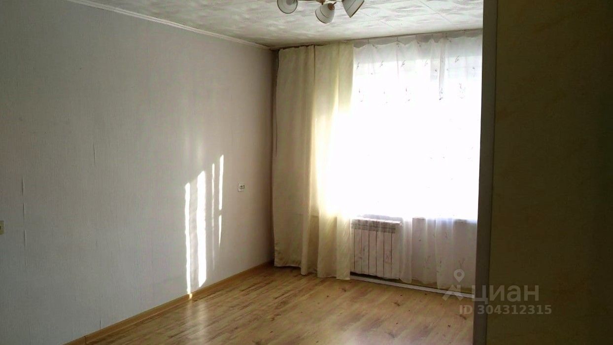 Внутреннее пространство оформлено в светлых тонах с акцентом на простоту и функциональность. Стены окрашены в нежный серый цвет, а на полу уложен светлый ламинат. Окно, обрамленное легкими занавесками, пропускает много естественного света, создавая атмосферу уюта. В комнате присутствует радиатор отопления под окном. Потолок оформлен с использованием простого покрытия, а освещение обеспечивается потолочной люстрой с несколькими источниками света. Общая планировка комнаты позволяет легко организовать пространство для различных нужд.