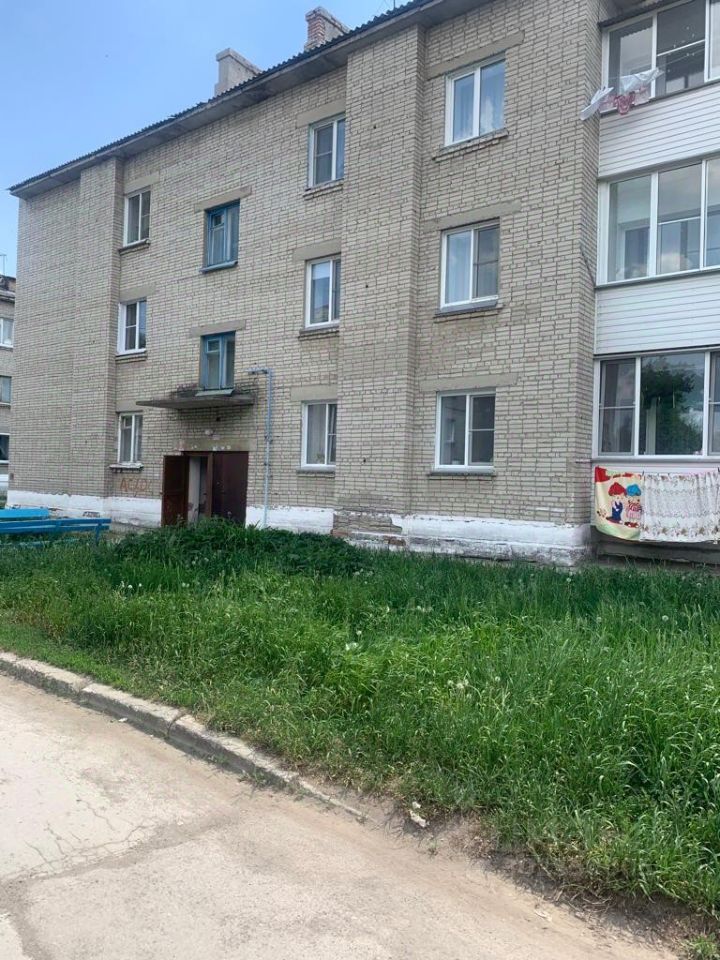 Здание выполнено в простом кирпичном архитектурном стиле с прямыми линиями и симметрией. Фасад имеет светло-коричневый цвет с крупными окнами, оформленными в белых рамках. Входная дверь расположена в центре, с небольшим козырьком. На первом этаже видна часть балкона с ярким текстилем. Окна второго этажа также имеют белые рамы. Перед зданием находится зеленая территория с высокой травой, создающей естественный контраст с кирпичной стеной. Общая отделка выглядит простой и функциональной, без излишеств.