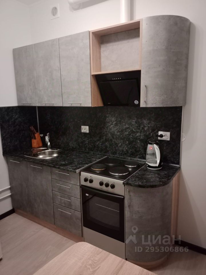 Кухня выполнена в современном стиле с элементами минимализма. Стеновые шкафы имеют бетонный и деревянный отделки, создавая гармоничное сочетание. Рабочая поверхность из темного камня, на которой расположены плита и раковина. Встроенная техника включает духовой шкаф и вытяжку, что подчеркивает функциональность пространства. Цветовая гамма включает серые и черные тона, что придает интерьеру стильный и современный вид. Основные элементы дизайна — гладкие линии и округлые углы, создающие уютную атмосферу. Пространство организовано эффективно, что позволяет удобно использовать кухонные принадлежности.