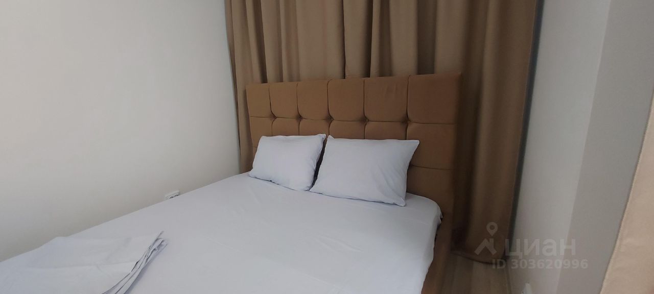 Уютная спальня с мягкой кроватью и белыми подушками, светлые стены, коричневые шторы, аккуратная и спокойная атмосфера