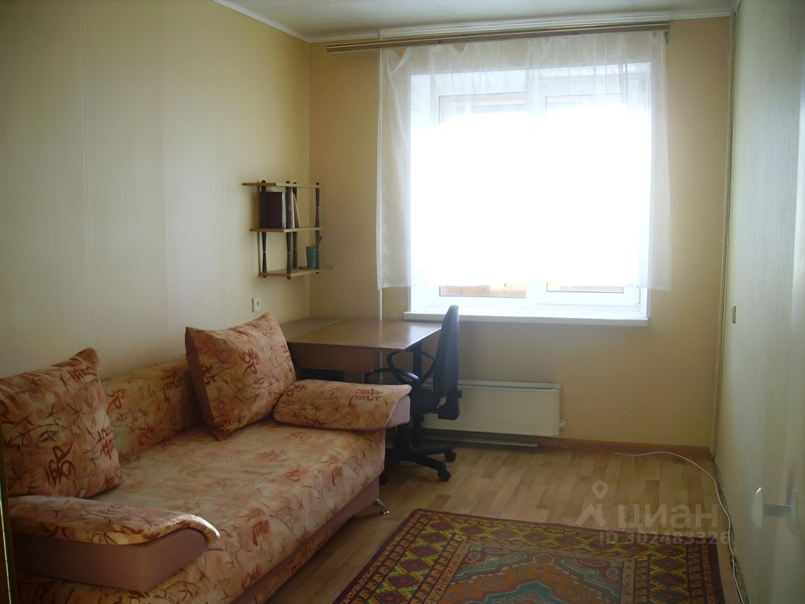 Светлая комната с диваном и рабочим столом, окно с тюлем, уютная атмосфера, деревянный пол, ковёр, полки для книг
