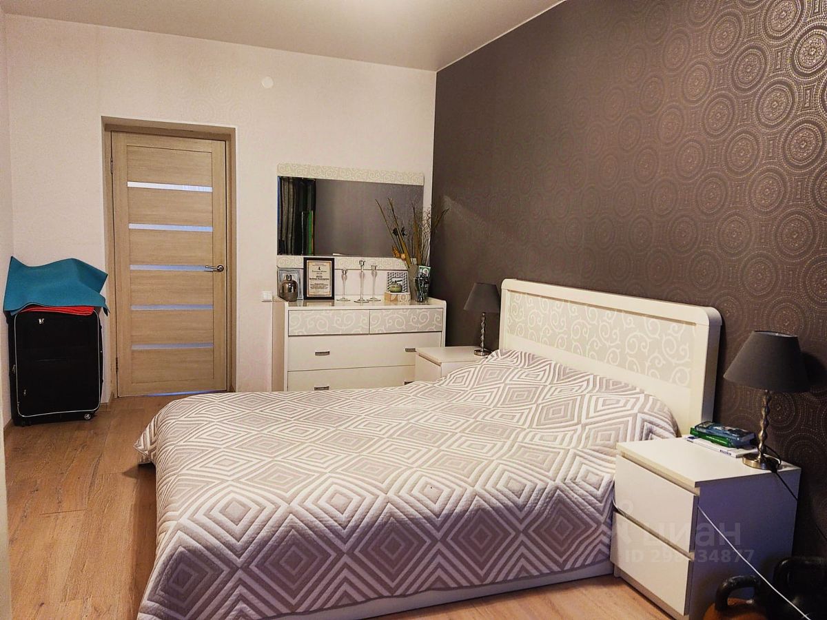 Спальня оформлена в современном стиле с акцентом на уют и функциональность. Стена с узорчатыми обоями темного цвета создает атмосферу спокойствия. Кровать с белым изголовьем и геометрическим покрывалом занимает центральное место. По обе стороны от кровати расположены прикроватные тумбочки с настольными лампами. На стене над тумбочками находится зеркало с декоративной рамкой. Дверь из светлого дерева с прозрачными вставками добавляет светлоты. Пол из ламината теплого оттенка придает комнате завершенный вид, а элементы декора, такие как цветы в вазе, подчеркивают стиль.