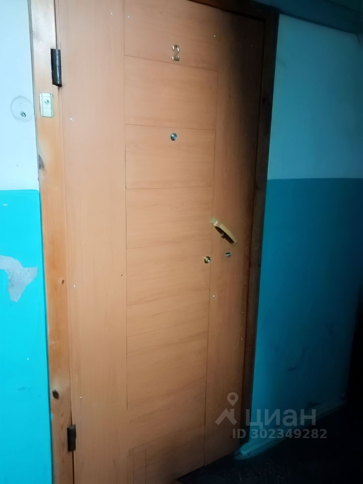 Дверь выполнена в современном стиле с использованием древесного материала, окрашенного в светлый оттенок. На двери расположены металлические элементы, включая замок и ручку. Номер квартиры обозначен в верхней части двери. Стены вокруг двери имеют двухцветное оформление: нижняя часть окрашена в яркий голубой цвет, а верхняя — в нейтральный светлый оттенок. Пол выполнен из темного материала, что создает контраст с яркими стенами. Общий вид пространства создает ощущение простоты и функциональности.