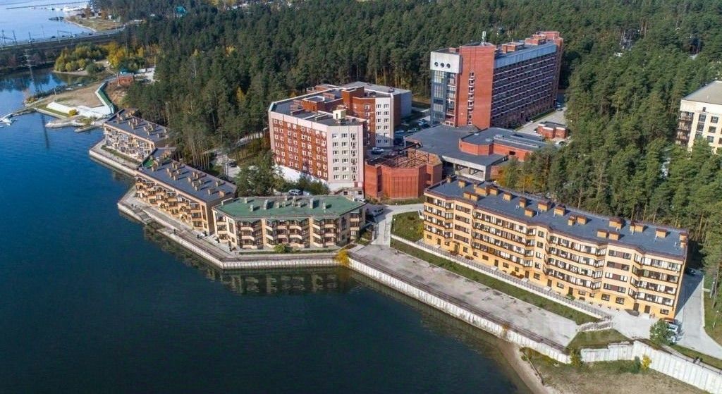 Современные здания в стиле минимализма с прямыми линиями и большими окнами. Видны многоквартирные дома с террасами и балконами, выполненные из кирпича и бетона. Структурные элементы включают плоские крыши и обширные фасады. Здания расположены вдоль водоема, создавая гармоничное сочетание с природой. Цветовая палитра включает теплые оттенки кирпича и нейтральные тона. Отделка фасадов из современных материалов придает им стильный и ухоженный вид. Пространство организовано для комфортного проживания, с акцентом на функциональность и эстетику.