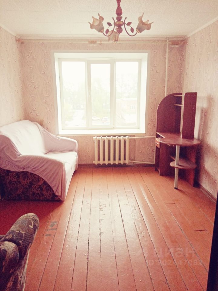 Комната оформлена в простом стиле с обоями с рельефным узором. На полу находятся деревянные полы, окрашенные в теплый оранжевый цвет. У окна расположено радиаторное отопление, а также стоит диван с чехлом. Угловой письменный стол из темного дерева дополнен полкой. Центральное освещение представлено люстрой с несколькими лампами. Окно обеспечивает естественное освещение, создавая уютную атмосферу. Стены комнаты светлые, что визуально увеличивает пространство.