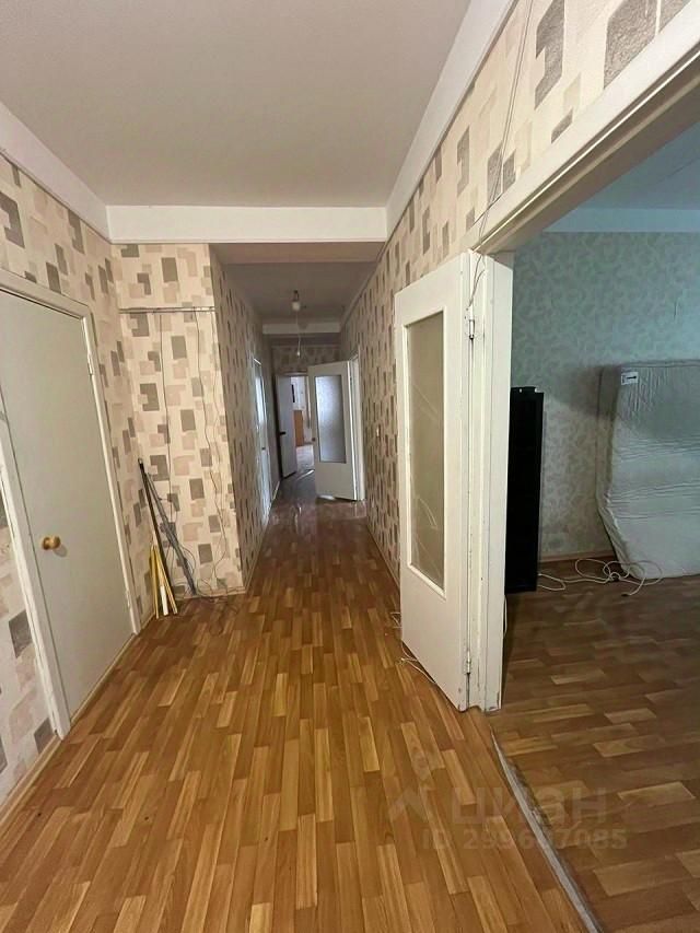 Видна длинная прихожая с деревянным полом и обоями в светлых тонах с геометрическим узором. Стены оформлены в нейтральных цветах, создавая ощущение простора. На полу уложен паркет, что добавляет тепла и уюта. Двери с прозрачными вставками из стекла ведут в соседние комнаты. Освещение мягкое, потолок белый, что визуально увеличивает пространство. В конце коридора видна другая комната с мебелью, частично скрытой от взгляда. Общая атмосфера помещения спокойная и современная, что делает его привлекательным для проживания.