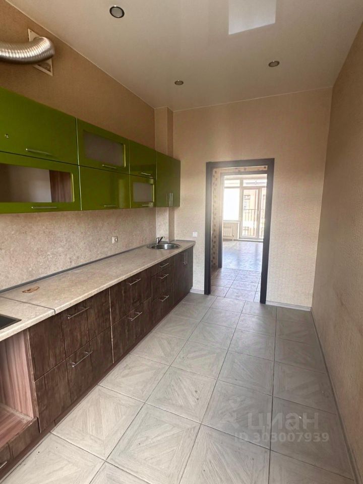 Кухня оформлена в современном стиле с элементами минимализма. Верхние шкафчики яркого зеленого цвета контрастируют с темной древесиной нижних ящиков. Рабочая поверхность выполнена из светлого материала, что создает ощущение простора. Видно, что в помещении установлена раковина и имеется место для встраиваемой техники. Пол выложен плиткой с геометрическим узором, что добавляет текстуру. Стены отделаны светлым материалом, создающим уютную атмосферу. Дверной проем ведет в другую зону, что позволяет естественному свету проникать в кухню.