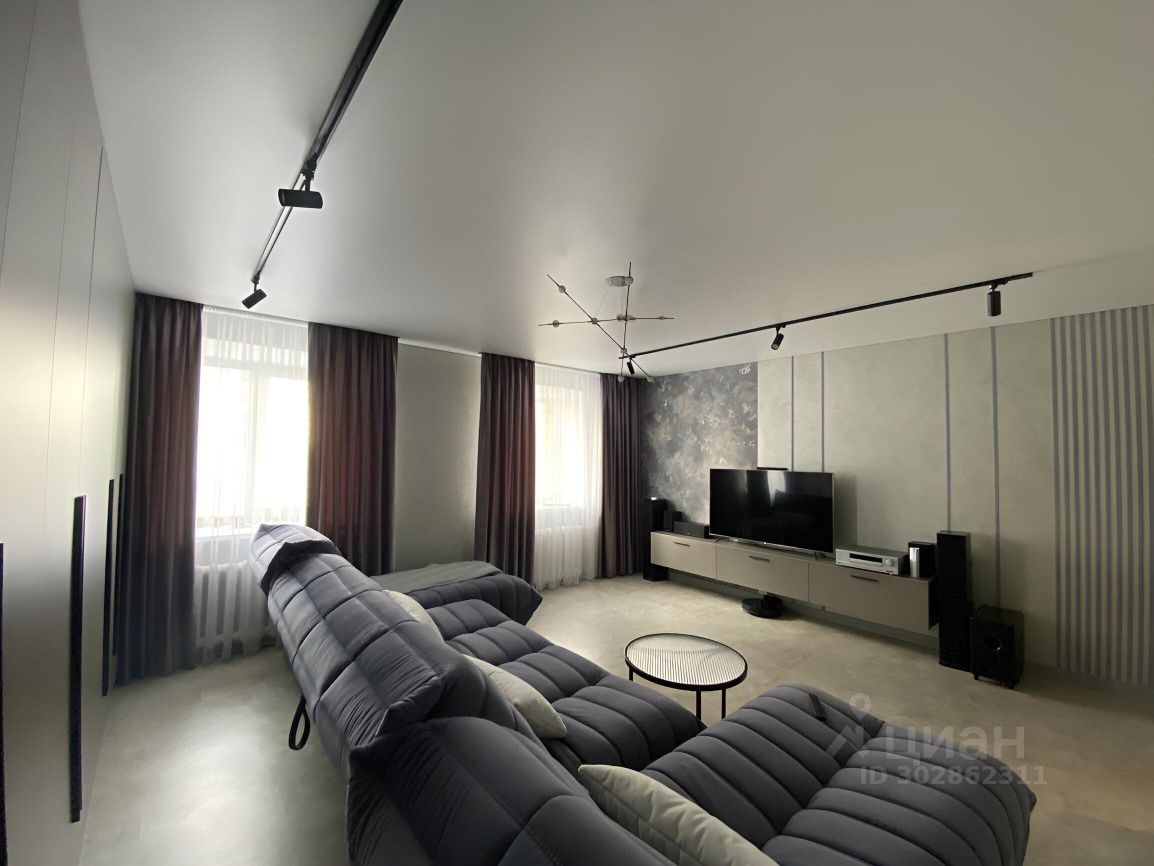 Современный интерьер с элементами минимализма. Просторная гостиная с мягким угловым диваном серого цвета, который создает уютную атмосферу. На стене расположен телевизор, а под ним — стильный низкий шкаф с техникой. Окна обрамлены длинными шторами в темных тонах, что добавляет глубину пространству. Потолок с встраиваемыми светильниками и оригинальной люстрой создает мягкое освещение. Стены оформлены в нейтральных оттенках с текстурированными элементами, что придает комнате современный вид. Пол выполнен из светлого материала, что визуально расширяет пространство.