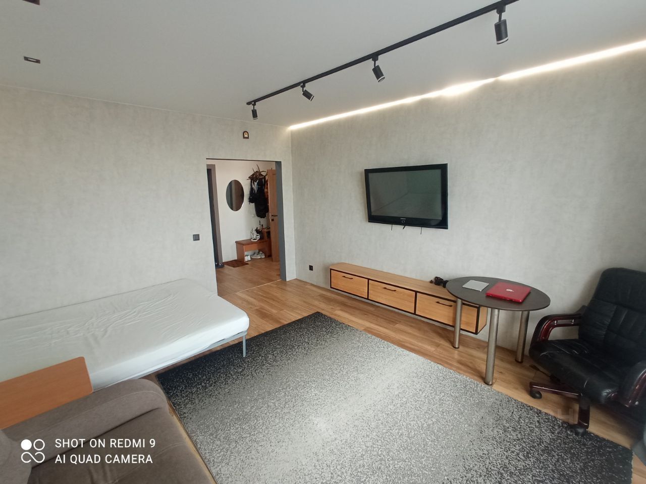 Комната оформлена в современном стиле с нейтральными цветами стен и деревянным полом. На стене установлен плоский телевизор, под которым располагается низкий комод с выдвижными ящиками. В углу стоит круглый стол с офисным креслом, что создает рабочую зону. В комнате видна часть дивана с матрасом, что указывает на возможность использования пространства как спальни. Освещение представлено в виде встроенных светильников и светодиодной ленты, создающих уютную атмосферу. Общая отделка выглядит аккуратно и стильно, что подчеркивает функциональность и комфорт помещения.