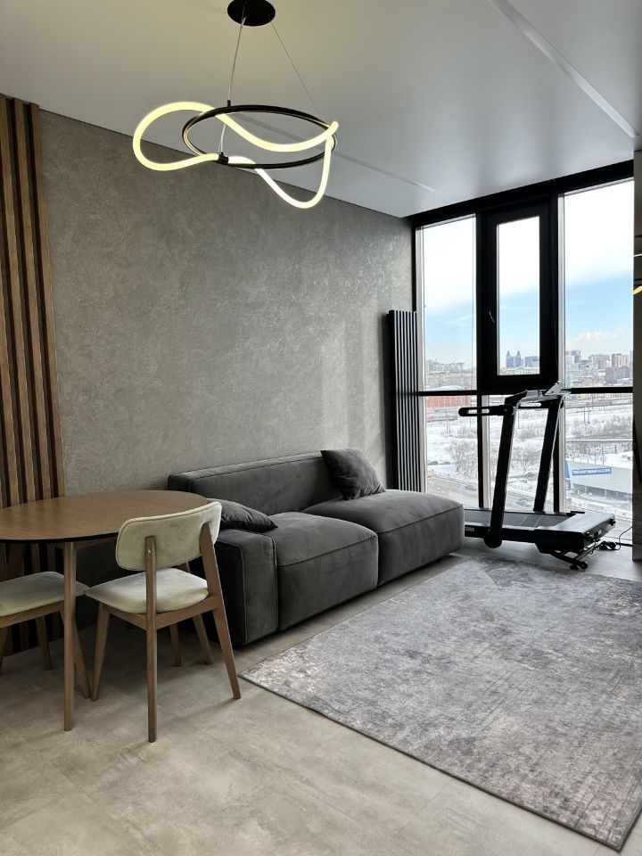 Современный интерьер с минималистичным дизайном. Стена отделана серым текстурированным материалом, дополненная деревянными панелями. В комнате расположены уютный серый диван и круглый стол из дерева с четырьмя стульями, один из которых обит мягким материалом. Пол покрыт светлой плиткой, а в центре находится большой ковер с абстрактным узором. Освещение представлено стильной люстрой с изогнутыми линиями. Окна от пола до потолка обеспечивают обилие естественного света и открывают вид на город.
