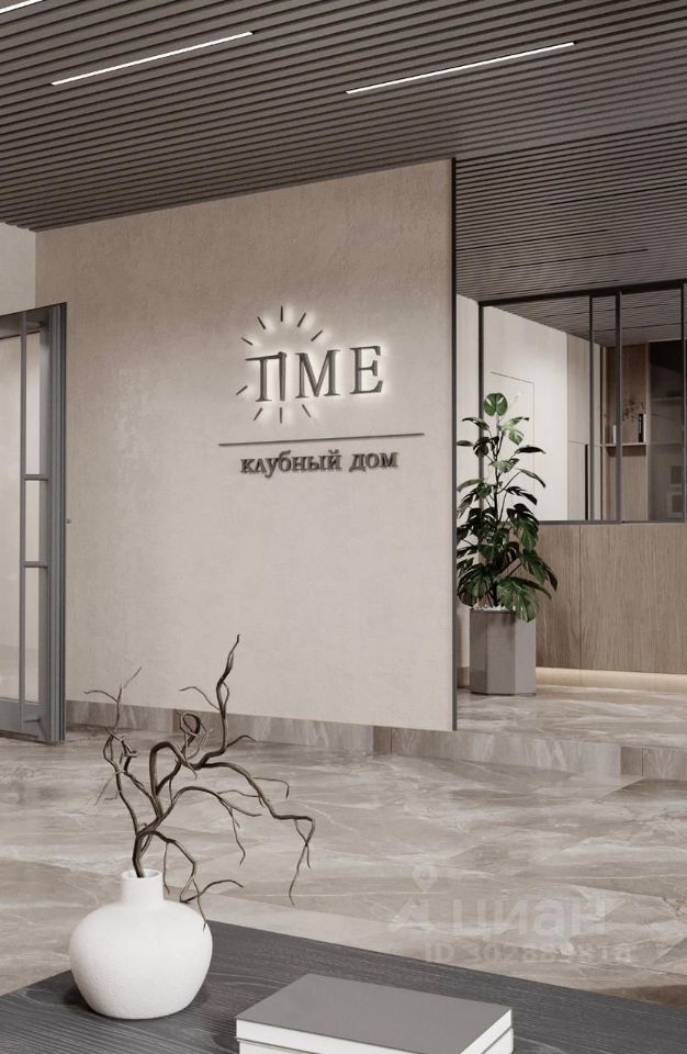 Современный интерьер с минималистичным дизайном, выполненный в светлых тонах. Стены отделаны гладкой штукатуркой, а потолок украшен деревянными панелями с подсветкой. Основное пространство включает в себя зону с декоративными растениями и стильной мебелью. Пол покрыт блестящей плиткой, отражающей свет. На переднем плане находится стол с вазой и сухими ветками, что добавляет элегантности. Логотип на стене подчеркивает уникальность этого клуба, создавая атмосферу уюта и современности.