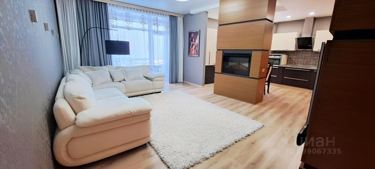 Современный интерьер с элементами минимализма. Просторная гостиная объединена с кухней, выделяется уютный белый диван и мягкий ковер. Стены отделаны светлыми тонами, с акцентами на текстурированной поверхности. Открытые окна с легкими занавесками обеспечивают естественное освещение. Кухонные шкафы выполнены в темных оттенках, создавая контраст с общим светлым фоном. Встроенный камин из дерева служит центральным элементом, добавляя тепла и уюта. Лампы с современным дизайном дополняют атмосферу, создавая гармоничное пространство для отдыха и общения.