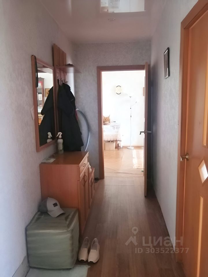 Коридор с простым и функциональным дизайном. Стены окрашены светлым цветом, создавая ощущение пространства. На стене имеется зеркало и вешалка для одежды, что добавляет удобства. Пол выложен ламинатом, а в углу стоит небольшая тумба с ящиками. На ней расположены бутылка с чистящим средством и кепка. У двери видны туфли, а также небольшой мягкий пуф. В конце коридора открывается вид на светлую комнату с мебелью, что добавляет уют в пространство.