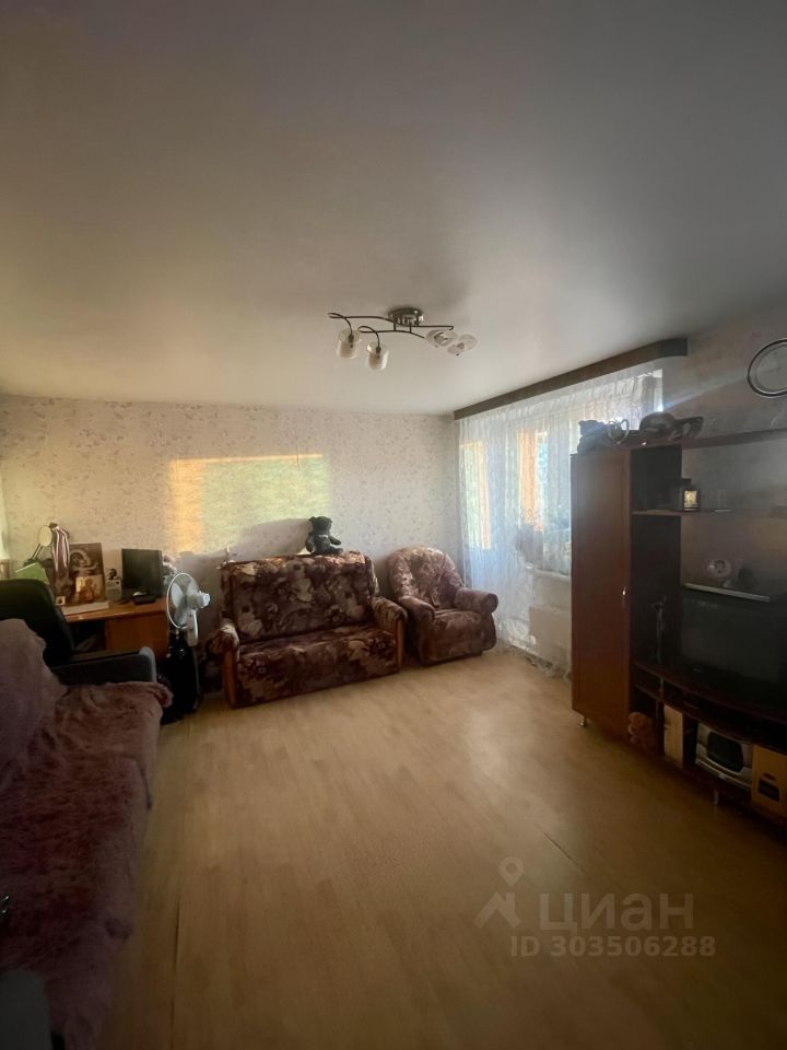 Комната оформлена в уютном стиле с мягкой мебелью, обитой тканью с цветочным принтом. На стенах светлые обои с ненавязчивым узором, потолок ровный и светлый. Основное освещение обеспечивается современным светильником с несколькими лампами. В комнате находятся два дивана и кресло, создающие комфортную зону для отдыха. Пол выполнен из светлого дерева, что добавляет тепла в интерьер. У окна расположены легкие занавески, позволяющие пропускать естественный свет. В углу стоит телевизор на тумбе, а рядом находится стол с предметами декора.