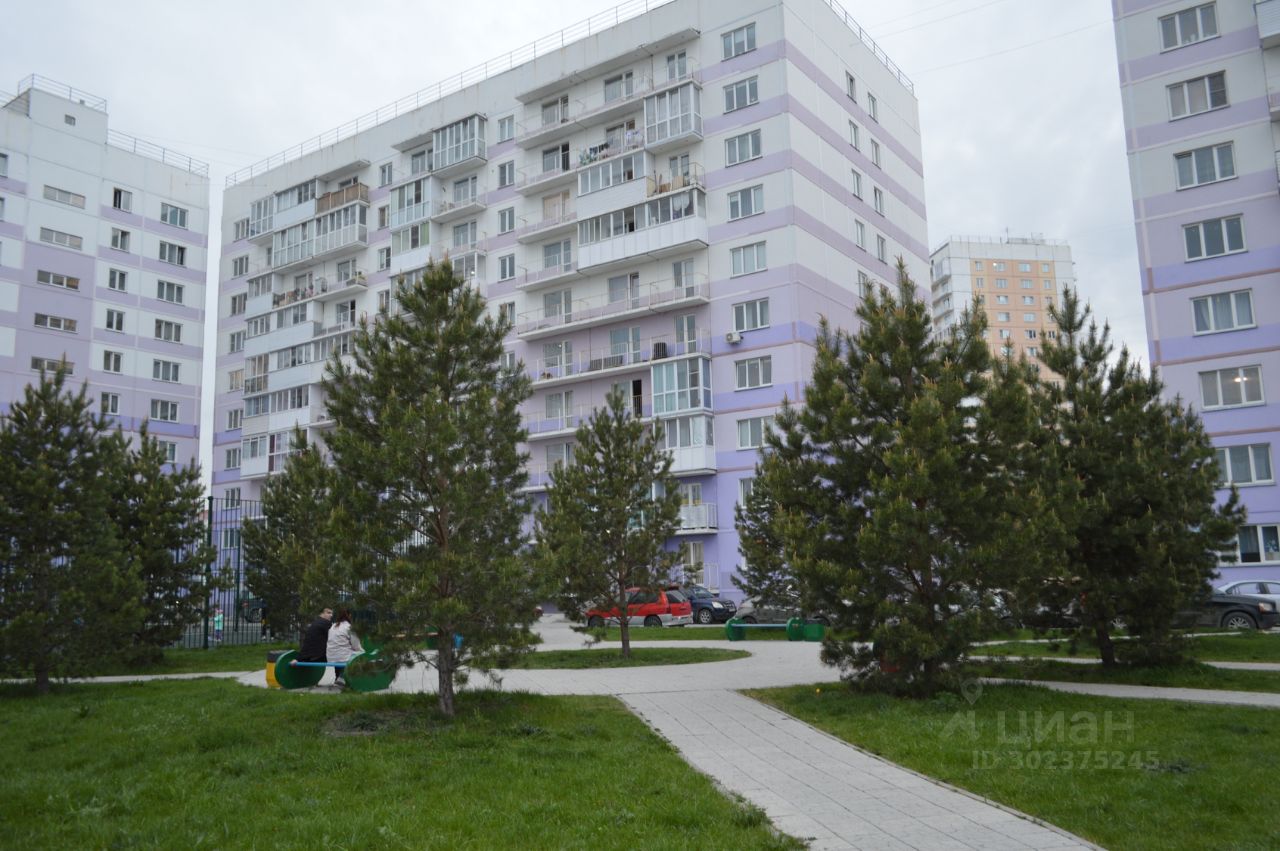 Многоэтажное здание выполнено в современном архитектурном стиле с элементами минимализма. Фасад окрашен в светлые тона с фиолетовыми вставками, что придает ему свежий вид. Видны балконы с остеклением, что добавляет функциональности. Внутренний двор оформлен зелеными насаждениями и тротуарной плиткой, создавая уютное пространство для отдыха. Основные материалы — бетон и стекло, а цветовая палитра включает белый, фиолетовый и зеленый. Общая эстетика здания и двора подчеркивает гармонию с окружающей средой и современный подход к жилому строительству.