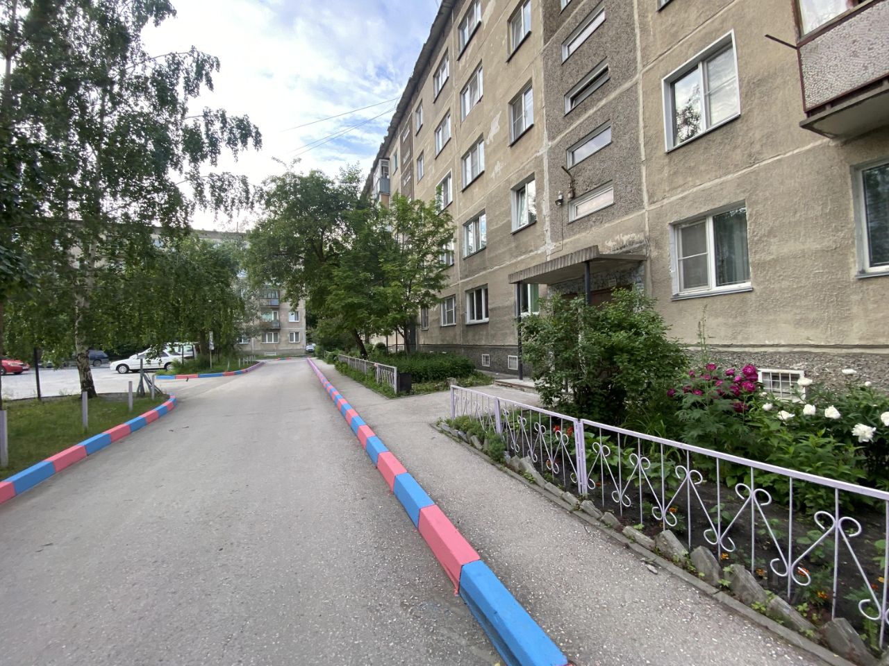 Здание выполнено в советском архитектурном стиле, с характерными серыми бетонными стенами и простыми формами. На фасаде видны окна с металлическими рамами и небольшими балконами. Входная зона оформлена крышей и небольшими зелеными насаждениями. Дорога перед зданием вымощена асфальтом, с яркими цветными бордюрами в розовых и голубых тонах. Участок вокруг здания озеленен, с кустарниками и цветами, что придаёт уютную атмосферу. Структурные элементы, такие как перила и бордюры, выполнены из металла и бетона, создавая гармоничный внешний вид.