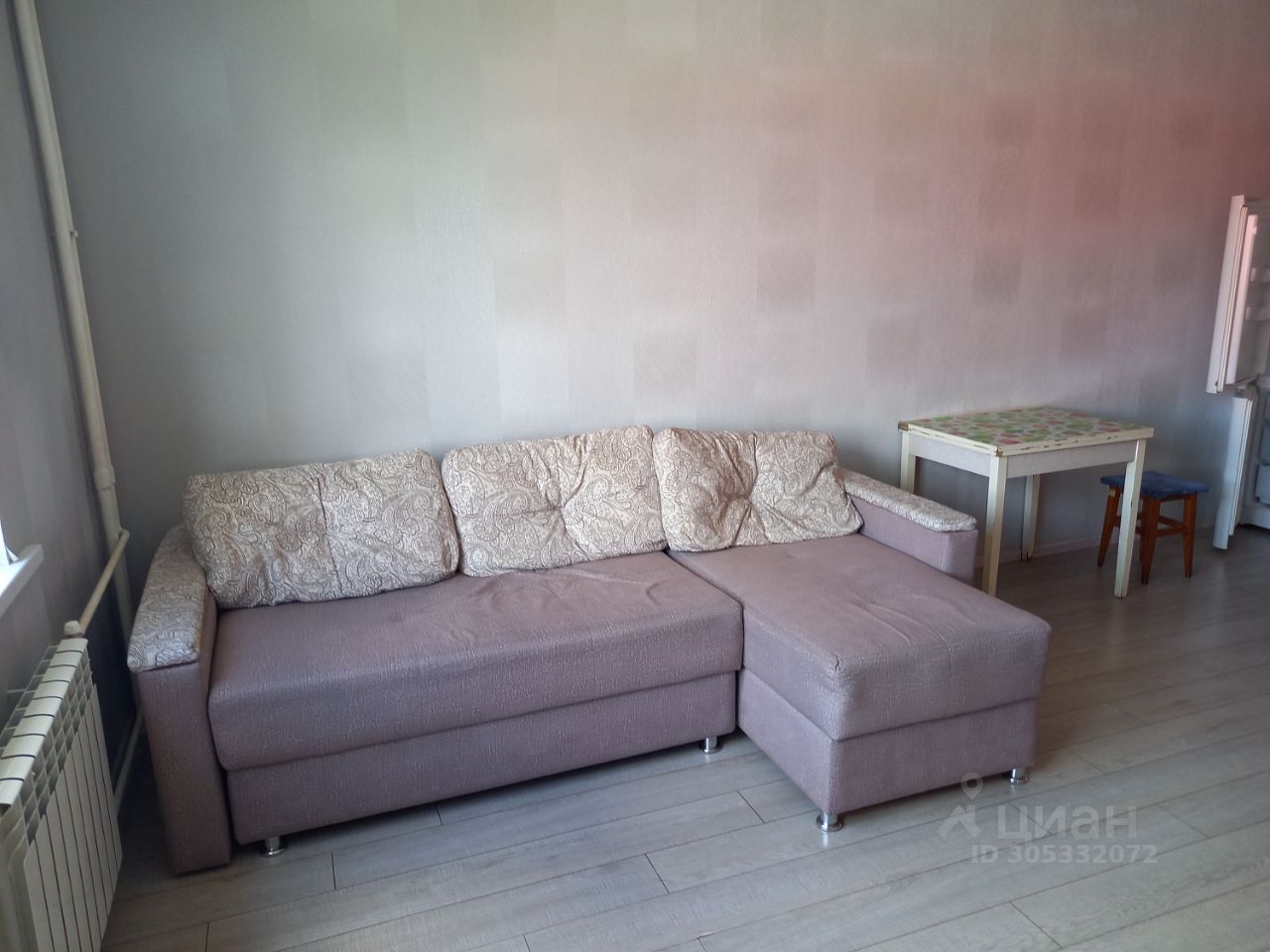 В комнате расположен угловой диван с текстурной обивкой, выполненный в светлых тонах. Стены окрашены в нежный серый цвет, создавая ощущение простора. На полу уложен светлый ламинат, что добавляет современности интерьеру. Рядом с диваном стоит небольшой стол с цветной столешницей и стул, выполненный из дерева. В углу виден радиатор отопления, обеспечивающий комфортную температуру. Пространство выглядит уютным и функциональным, идеально подходящим для отдыха или работы.