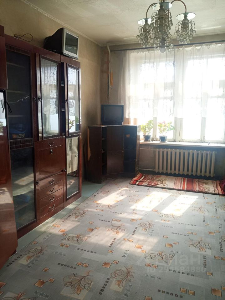Комната оформлена в классическом стиле с элементами советского дизайна. На стенах светлые обои, а на полу расположен ковролин с цветочным узором. В углу стоит темный деревянный шкаф с зеркальными дверцами и встроенным телевизором. Рядом находится небольшой стеллаж с телевизором и цветочными горшками на подоконнике. Окно обрамлено легкими занавесками, пропускающими свет. Потолок украшен хрустальной люстрой, создающей уютную атмосферу. Открытое пространство комнаты делает её светлой и комфортной для отдыха.