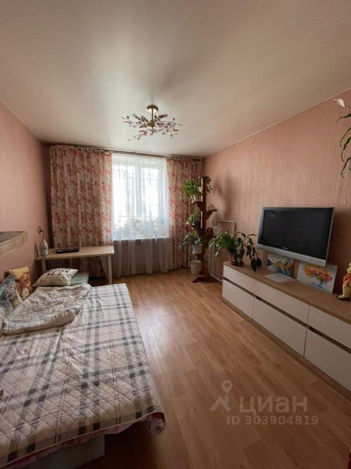Комната оформлена в мягких пастельных тонах с цветными обоями и деревянным полом. Основное внимание привлекает диван с пледом и подушками, расположенный рядом с телевизором на белом комоде. Окно, обрамленное легкими занавесками с цветочным узором, наполняет пространство естественным светом. В углу находятся горшки с растениями, добавляющие уют и свежесть. Потолок украшен стильной люстрой, создающей атмосферу домашнего уюта. Общая композиция комнаты создает ощущение спокойствия и комфорта.