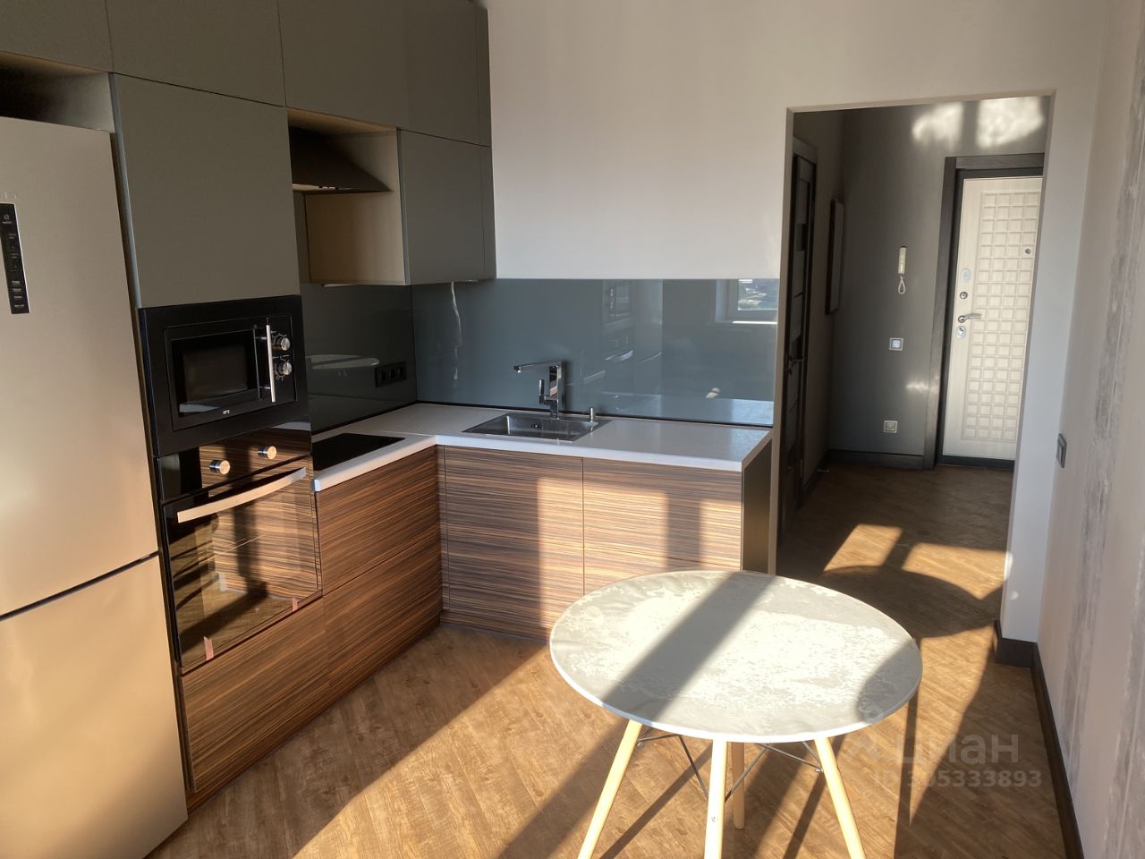 Кухня с современным дизайном, выполненная в стиле минимализм. Стеновые шкафы имеют гладкие серые фасады, а нижние шкафы отделаны текстурированным деревом. Встроенная техника включает холодильник и микроволновую печь, расположенные рядом с варочной панелью. Столешница белого цвета гармонирует с серой стеной и создает светлую атмосферу. Пол из светлого ламината добавляет теплоты пространству. В углу комнаты стоит круглый стол на тонких деревянных ножках, создающий уютное место для завтрака. Вход в коридор виден на заднем плане.