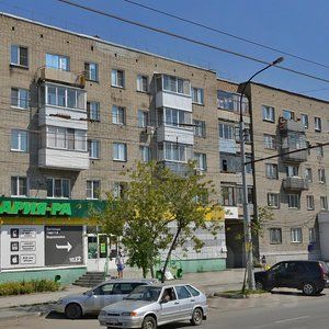 Фасад здания выполнен в советском архитектурном стиле с характерными элементами, такими как балконы и большие окна. Структура представляет собой многоквартирный дом с кирпичной отделкой, имеющий несколько этажей. На первом этаже расположены коммерческие помещения, включая магазин. Основные цвета фасада — серый и бежевый, что придаёт зданию строгий, но уютный вид. Балконы с металлическими перилами добавляют функциональности и визуального интереса. Пространство вокруг здания оформлено зелеными насаждениями, что создаёт приятную атмосферу для жителей и посетителей.