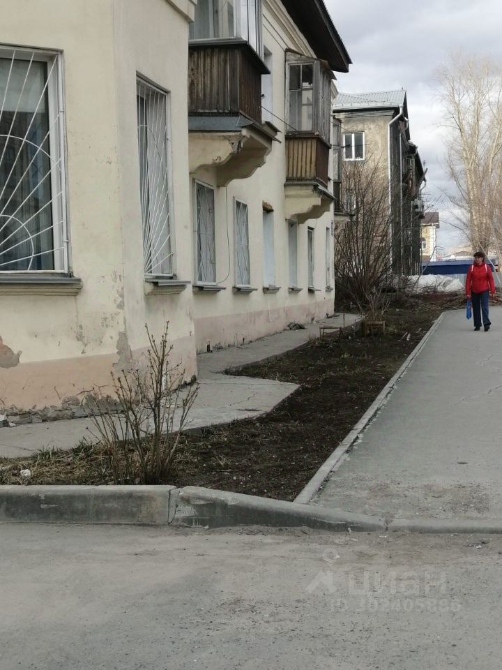 Здание выполнено в советском архитектурном стиле с простыми, но функциональными элементами. Видны большие окна с решетками, некоторые из которых имеют балконы. Стены здания окрашены в светлый цвет с частично обнаженной штукатуркой. На переднем плане расположена ухоженная территория с небольшими кустарниками и плиткой, ведущей к входу. Асфальтированная дорога проходит рядом, создавая доступ к зданию. Общий вид демонстрирует практичность и простоту, характерные для жилых домов этого периода.