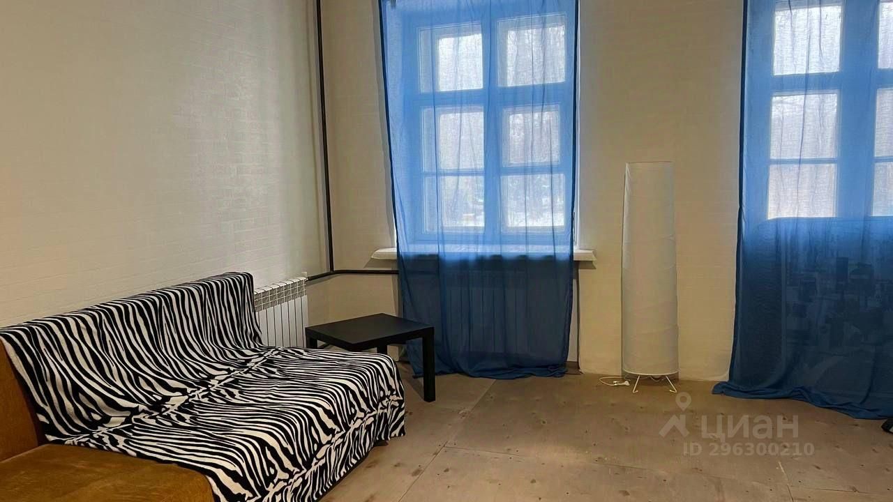 В комнате представлена современная обстановка с элементами минимализма. На переднем плане находится диван с зебровым принтом, создающий интересный акцент. Рядом расположен небольшой черный столик, обеспечивающий функциональность пространства. Окна украшены полупрозрачными синими занавесками, которые пропускают мягкий свет. Пол выполнен из светлого дерева, а стены окрашены в нейтральный цвет, что придаёт комнате ощущение простора и уюта. Отопительный радиатор добавляет практичности, обеспечивая комфорт в холодное время года.