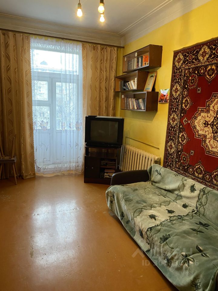 Комната оформлена в простом, но уютном стиле. На стенах желтая окраска, что придает пространству светлую атмосферу. В углу расположено окно с легкими занавесками, позволяющими естественному свету проникать внутрь. Удобный диван с покрывалом и подушками создает комфортную зону для отдыха. На стене висит большой ковер с традиционным узором, добавляющий тепла и текстуры. Пол выполнен из линолеума, который легко поддерживать в чистоте. Книжная полка с книгами и телевизор на тумбе дополняют функциональность пространства, создавая атмосферу домашнего уюта.