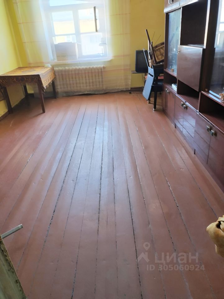 Комната с желтыми стенами и деревянным полом, выполненным в традиционном стиле. Пол имеет широкие доски, окрашенные в красный цвет. Окно с белыми рамами обеспечивает естественное освещение, а на подоконнике стоят небольшие предметы. В углу комнаты расположен шкаф с витринами, в который встроены стеклянные полки. Рядом стоит стол с резной отделкой, создающий уютную атмосферу. В комнате также виден радиатор отопления под окном. Общая обстановка выглядит простой и функциональной, с акцентом на деревянные и классические элементы интерьера.