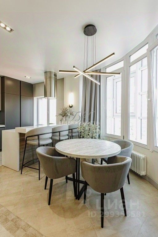 Современный интерьер столовой с круглым мраморным столом и мягкими серыми стульями. Стол окружен высокими стульями на металлических ножках, создающими стильный контраст. На потолке подвесная люстра с геометрическим дизайном, обеспечивающая мягкое освещение. Большие окна, обрамленные светлыми шторами, наполняют пространство естественным светом. Стены выполнены в нейтральных тонах, а пол покрыт плиткой с текстурой, что придает уют и элегантность. Кухонная зона, расположенная рядом, оформлена в темных тонах с современными фасадами и вытяжкой, создавая гармоничное сочетание стиля и функциональности.