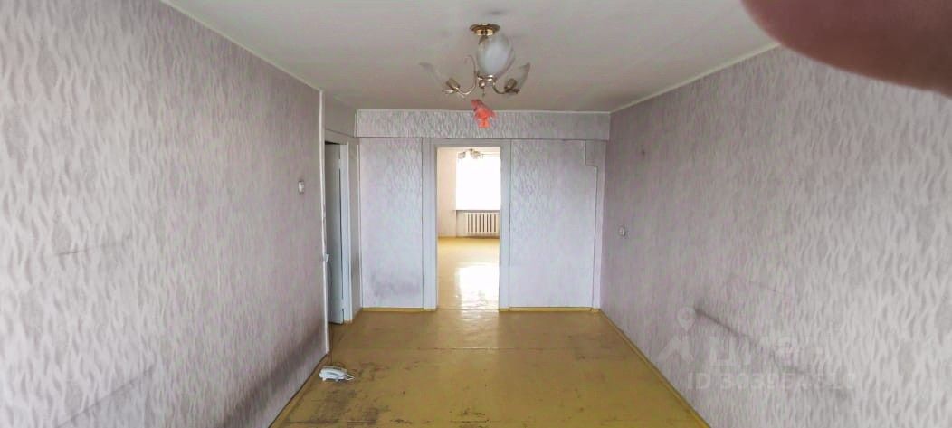 Видна комната с простым дизайном, оформленная в светлых тонах. Стены покрыты обоями с текстурным узором, создающим ощущение глубины. Пол выполнен из желтого линолеума, что придает помещению яркость. Центральная люстра обеспечивает общее освещение. Вход в соседнее помещение оформлен дверным проемом без двери, что открывает доступ к следующей комнате. Окно с решеткой на стене напротив позволяет естественному свету заполнять пространство, подчеркивая его простоту и функциональность. Общая атмосфера комнаты создает ощущение просторности и возможности для дальнейшей отделки.