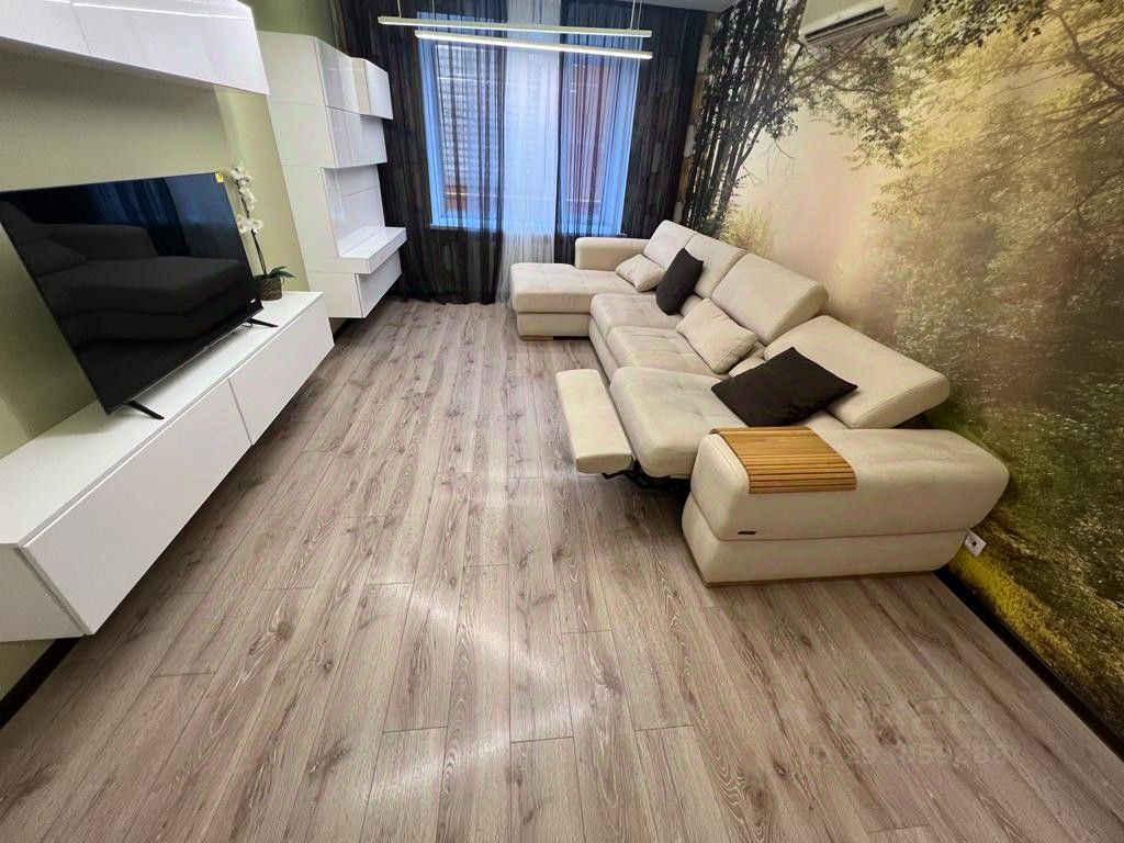 Современный интерьер с элементами минимализма. Просторная гостиная с угловым диваном светлого цвета и несколькими декоративными подушками. На стене изображен природный пейзаж, что создает уютную атмосферу. Напротив дивана расположена белая телевизионная тумба с плоским экраном. Полы выполнены из светлого дерева, что добавляет тепла в пространство. Освещение мягкое, с помощью потолочной лампы, создающей комфортное освещение. Окна закрыты полупрозрачными шторами, позволяя естественному свету проникать внутрь, при этом обеспечивая уединение.