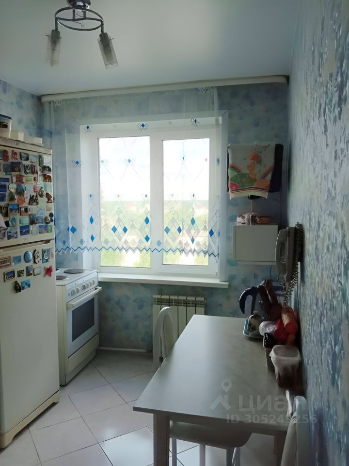 Кухня оформлена в светлых тонах с акцентами голубого цвета на стенах. На стенах расположены обои с легким узором. В комнате присутствуют большие окна с декоративными занавесками, пропускающими много света. Установлена белая газовая плита и холодильник, рядом стоит стол с двумя стульями. Пол выложен плиткой, а отопительный радиатор находится под окном. Пространство выглядит уютным и функциональным, идеально подходящим для приготовления пищи и семейных встреч.