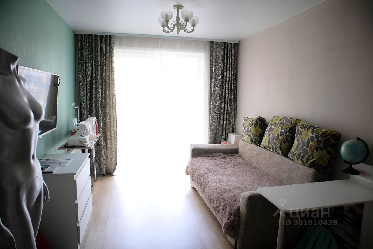 В комнате преобладает современный стиль с элементами минимализма. Стены окрашены в светлые тона, одна из стен имеет зеленый оттенок. Виден диван с мягким покрытием и декоративными подушками с цветочным рисунком. Окно, обрамленное длинными шторами с узором, обеспечивает естественное освещение. На стене установлен телевизор, рядом расположены швейные машинки на столе. Пол покрыт светлым ламинатом, создающим ощущение простора. Потолок украшен стильной люстрой, добавляющей уют в атмосферу комнаты.