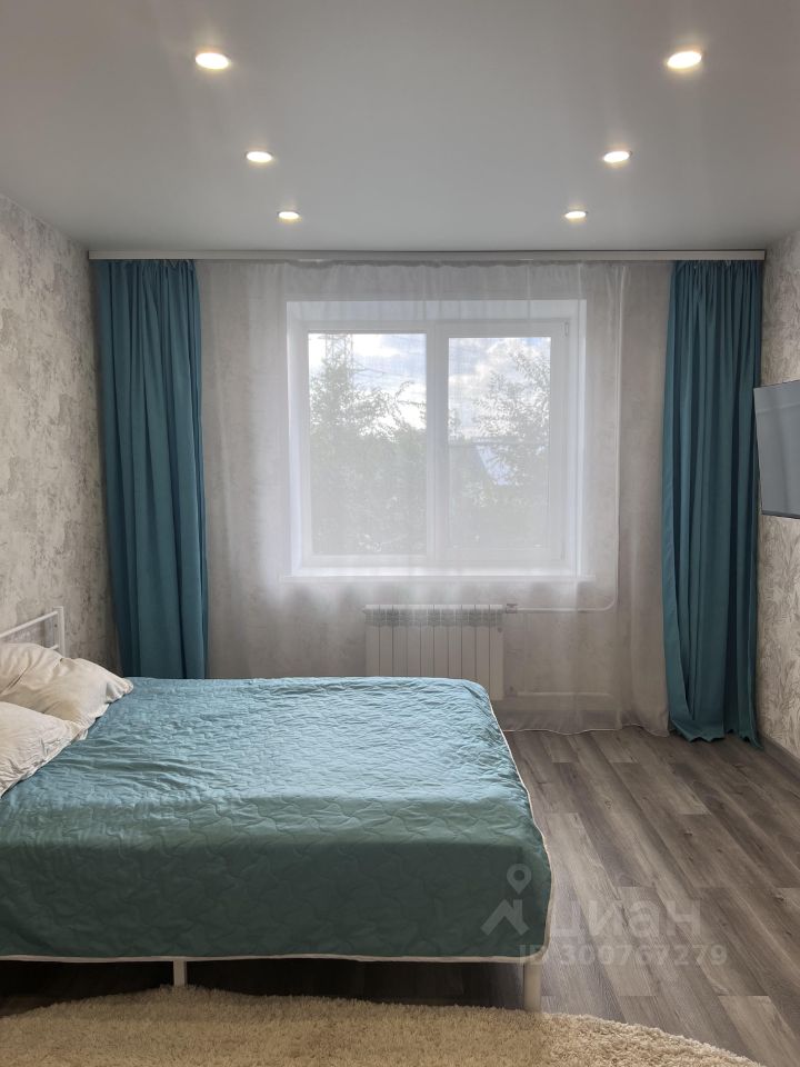 Спальня оформлена в современном стиле с акцентом на минимализм. Стены отделаны светлыми обоями с текстурой, создающими уютную атмосферу. Основной элемент интерьера — кровать с голубым покрывалом и двумя подушками, расположенная у стены. Окно с прозрачной занавеской и яркими шторами обеспечивает естественное освещение. Пол выполнен из светлого ламината, что добавляет пространству легкости. В комнате также присутствует радиатор отопления под окном и телевизор на стене, что делает пространство функциональным и комфортным для отдыха.