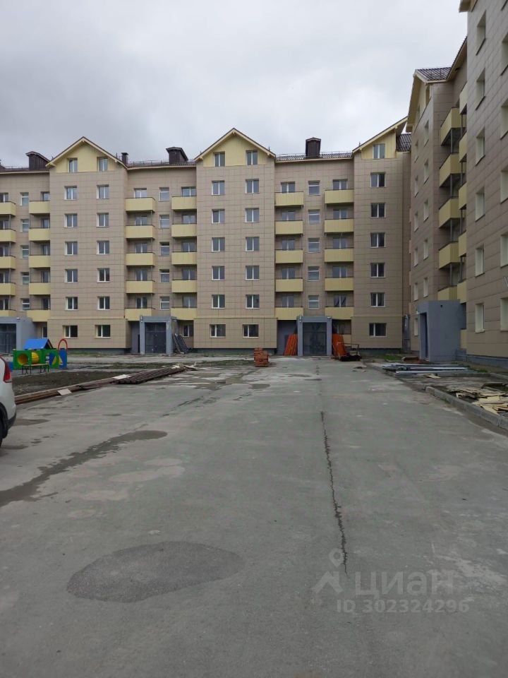 Здание выполнено в современном архитектурном стиле с элементами минимализма. Фасад облицован светло-бежевыми плитками, окна имеют прямоугольную форму и оформлены светлыми рамами. На каждом этаже расположены балконы с металлическими перилами, что добавляет легкости и открытости. Внутренний двор имеет асфальтированную дорожку, по обеим сторонам которой расположены входные группы с серыми дверями. Видна зона для детской площадки с игровыми элементами. Основные цвета — бежевый и серый, создают спокойную атмосферу. Общий вид подчеркивает функциональность и современность жилого комплекса.