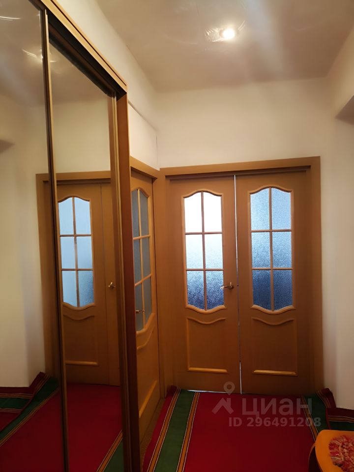 Видна прихожая с современным дизайном, где преобладают теплые оттенки. На стенах расположены двери с матовым стеклом и классическими ручками. Пол покрыт ярким ковром с зелеными и красными полосами, создающим уютную атмосферу. В одной из стен установлен зеркальный шкаф, который визуально расширяет пространство. Освещение представлено потолочным светильником, обеспечивающим достаточную яркость. Общий стиль интерьера подчеркивает гармонию между функциональностью и эстетикой.