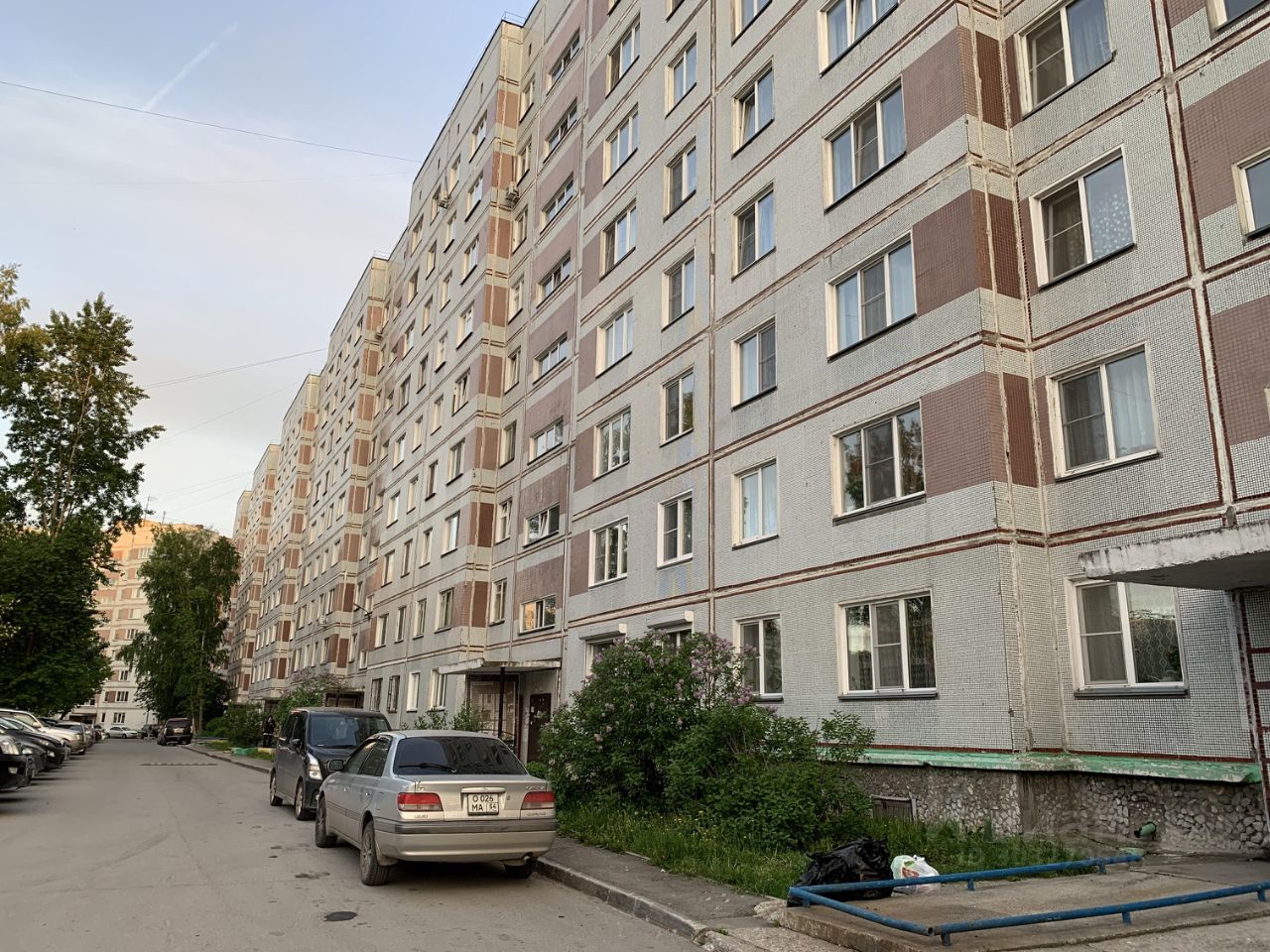 Здание выполнено в типичном советском архитектурном стиле, с характерной панельной конструкцией. Фасад украшен плиткой серого и коричневого цветов, а также горизонтальными линиями, создающими визуальную структуру. Окна имеют простые рамы и расположены симметрично, что придаёт фасаду упорядоченный вид. На первом этаже видно вход с крытой зоной, оформленный в минималистичном стиле. Вокруг здания растут кустарники и небольшие деревья, что добавляет зелени к общему облику. Асфальтированная парковка с автомобилями находится рядом, создавая функциональное пространство для жильцов.