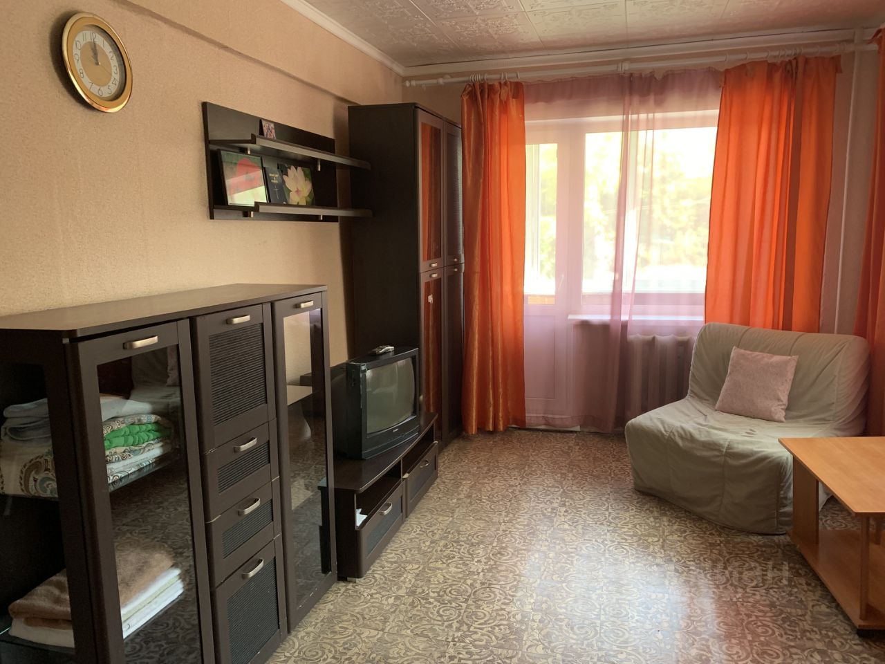 Комната оформлена в простом, функциональном стиле с элементами минимализма. На стенах светлые обои, а на полу находится узорчатый линолеум. Основные предметы мебели включают темный шкаф с прозрачными дверцами, телевизор и небольшой столик. Окна обрамлены яркими оранжевыми занавесками, создающими уютную атмосферу. В углу стоит мягкий диван с подушкой, что добавляет комфорт. Полки на стене украшены фотографиями, что придаёт пространству личный характер. В целом, комната выглядит светлой и удобной для отдыха.