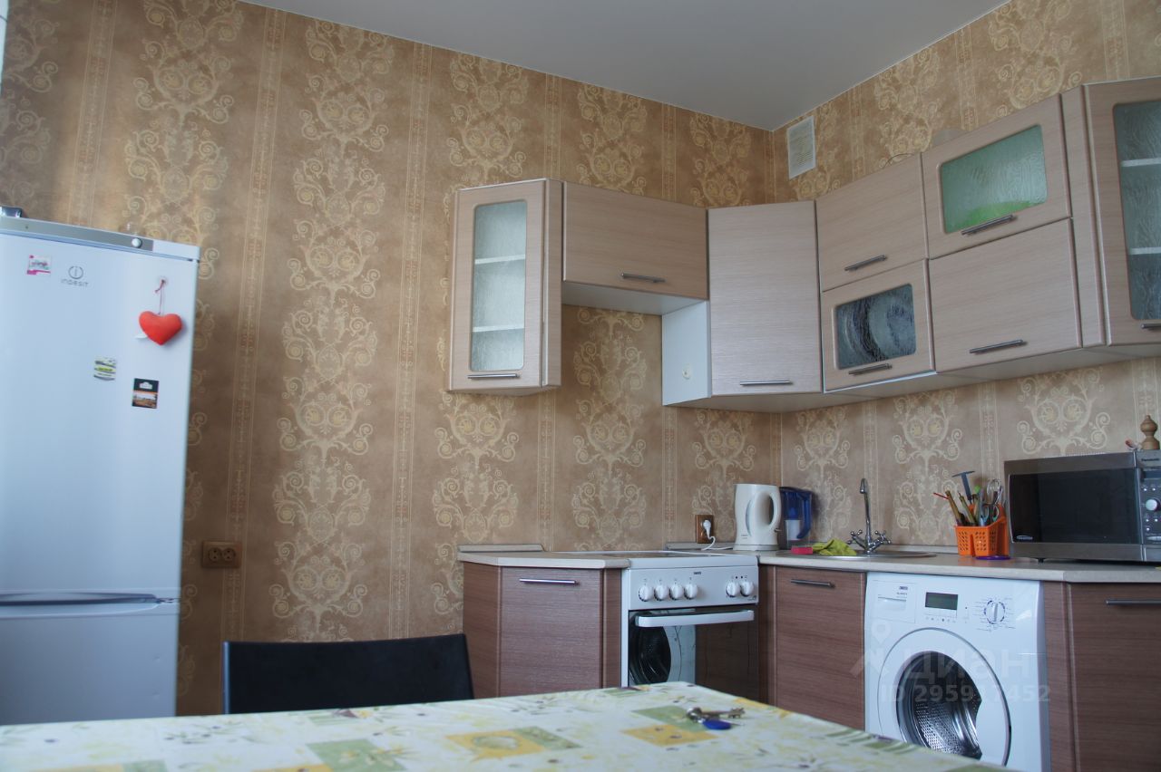 Кухня оформлена в современном стиле с элементами минимализма. Стены покрыты обоями с изысканным узором в теплых тонах. Встроенные шкафы из светлого дерева гармонично сочетаются с темной столешницей. На кухне расположены основные бытовые приборы: холодильник, плита, микроволновая печь и стиральная машина. Стол накрыт скатертью с ярким принтом, создающим уютную атмосферу. В углу виден чайник и контейнер для кухонных принадлежностей. Пространство выглядит функционально и комфортно, идеально подходя для повседневного использования.