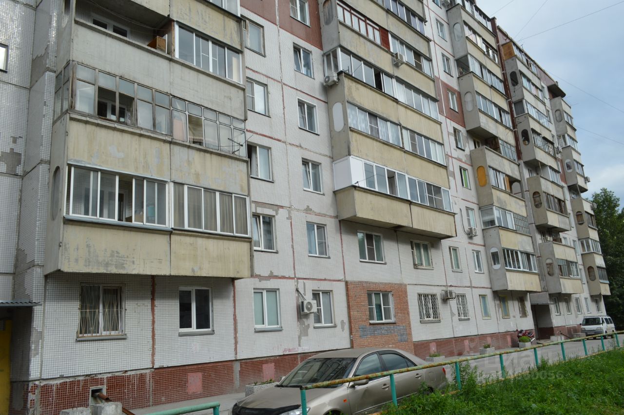Архитектурный стиль здания представляет собой типичную панельную конструкцию с элементами советского модернизма. Структурные особенности включают балконы с остеклением, а также разнообразные окна, некоторые из которых имеют округлую форму. Внешняя отделка выполнена из плитки и окрашенного бетона, что создает контраст между различными уровнями. Цветовая палитра включает светлые и темные оттенки, а также элементы кирпичной кладки. Финишные материалы выглядят изношенными, что указывает на возраст здания и необходимость ремонта.