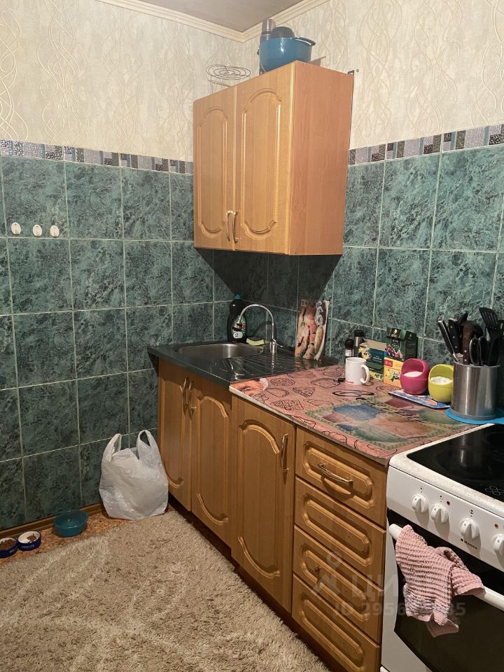 Кухня в простом стиле с элементами функционального дизайна. На стенах видна плитка зеленого цвета, а верхние шкафчики выполнены из светлого дерева. Рабочая поверхность покрыта яркой скатертью с рисунком. В углу расположена мойка с металлической раковиной и несколькими кухонными принадлежностями. Пол устлан ковриком, создающим уютную атмосферу. На столе находятся посуда и аксессуары, включая пластиковые чашки и контейнеры. Общая палитра включает зеленые и древесные оттенки, что придает пространству теплый и домашний вид.