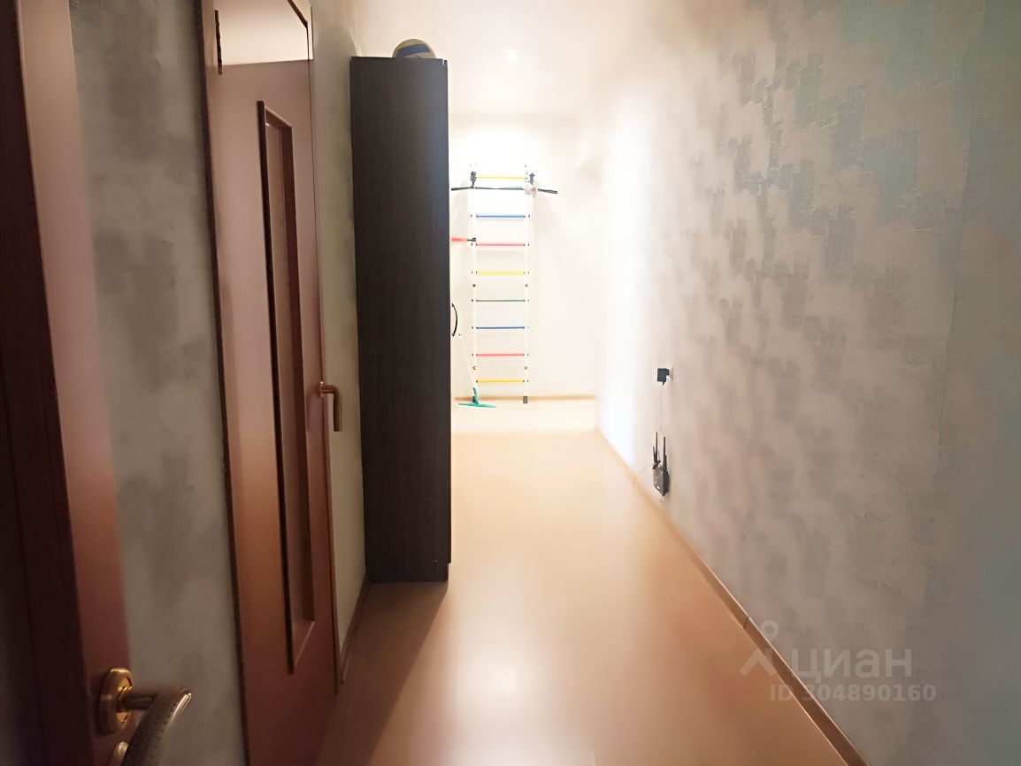 Длинный коридор с современным дизайном, отделанным светлыми стенами с текстурой. Слева расположены двери, выполненные в темном древесном цвете, что создает контраст с общей светлой палитрой. На одной из стен виден шкаф, также темного цвета. Пол коридора выполнен из гладкого материала, отражающего свет, что визуально увеличивает пространство. Вдалеке виден светлый участок, возможно, ведущий в другую комнату, и спортивная конструкция, добавляющая функциональности. Общая атмосфера пространства — современная и уютная.